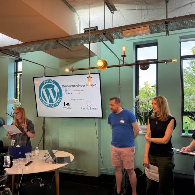 A woman stands at a table with a laptop and microphone, reading from a sheet of paper. Three people stand nearby, two holding papers, in front of a screen displaying the 