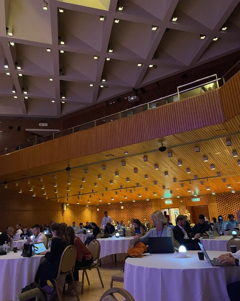 A large conference room with round tables, where people are seated using laptops and tablets under a geometric ceiling and warm lighting.