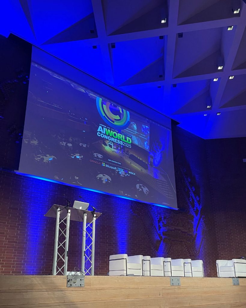 A stage with a podium and white, modular armchairs are set up in front of a large screen displaying 