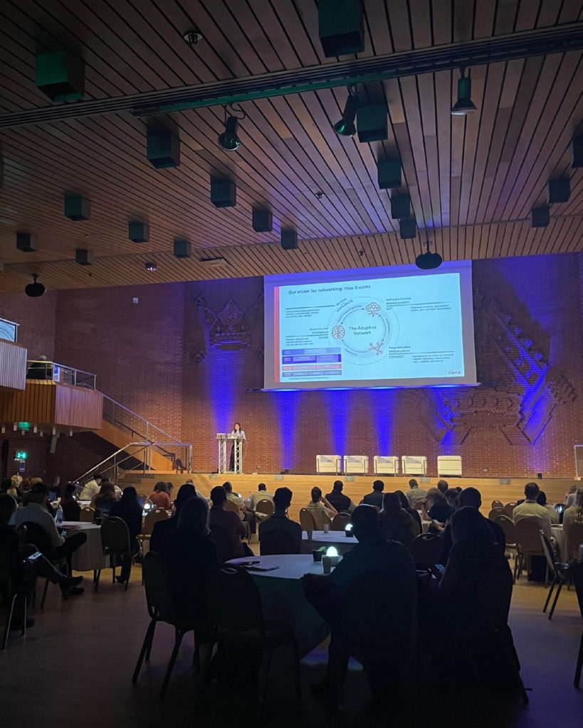A conference hall with people seated at round tables, watching a speaker present slides on a large screen in front of a brick wall.