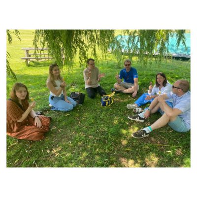A group of six people sit in a circle on grass under a leafy tree in a park, enjoying ice creams. Picnic tables and an open field is visible in the background.