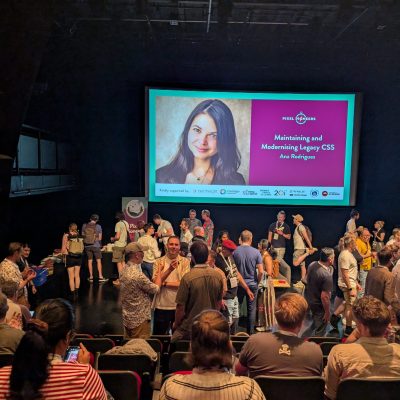 A conference auditorium with people standing and talking in front of a large screen displaying a presentation titled 